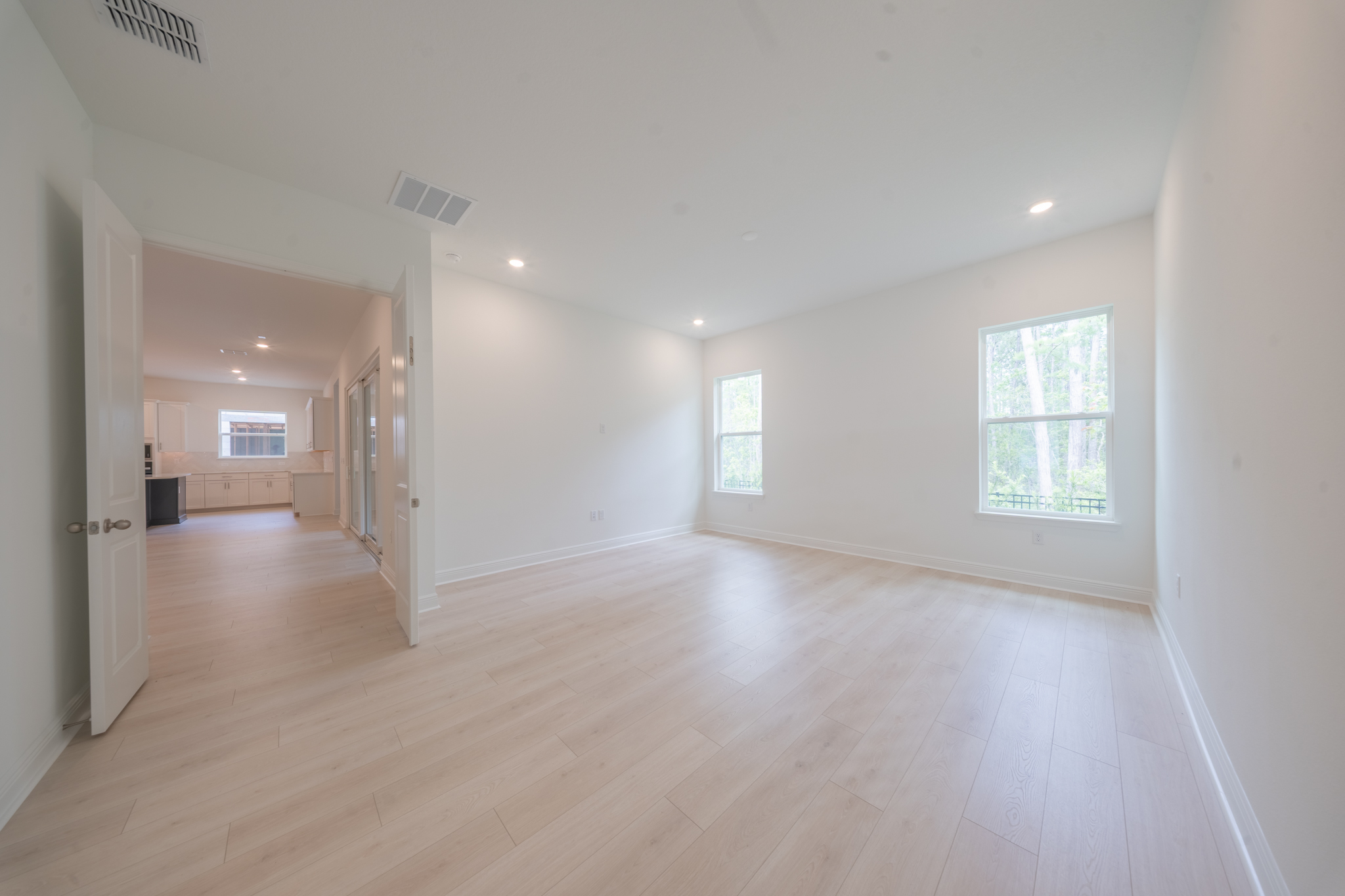 Empty primary bedroom with light hardwood floors and two windows overlooking trees