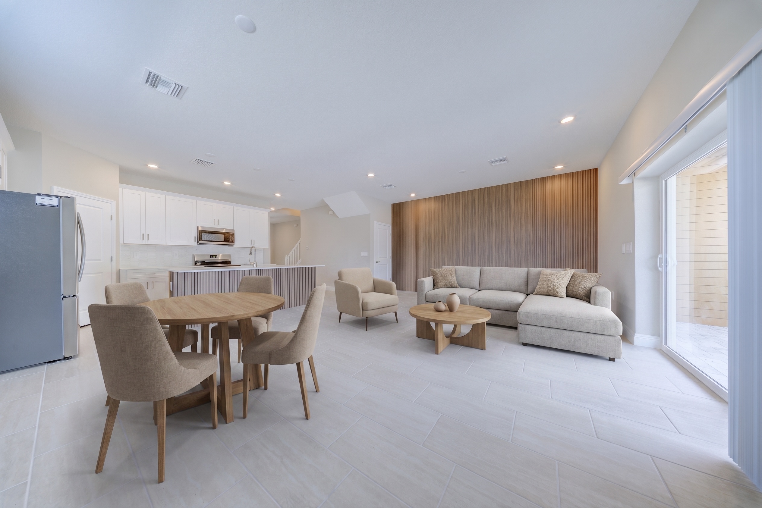 Virtually staged open-plan living and dining with round wood dining table, neutral sectional sofa and kitchen in background
