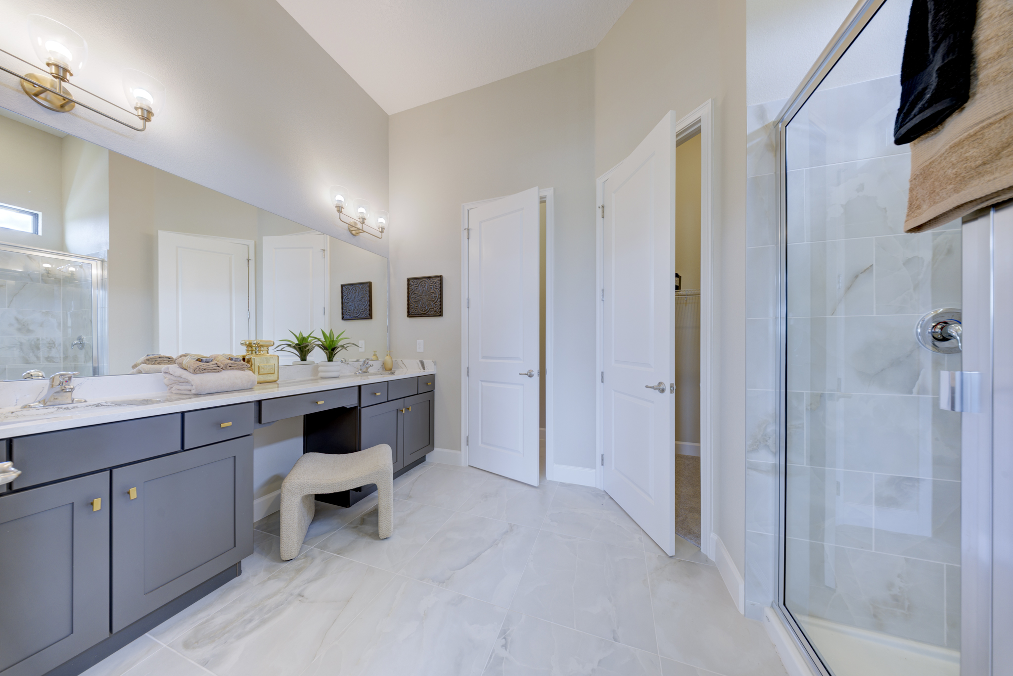 Primary bathroom with double gray vanity, marble tile floor, gold sconces and frameless glass walk-in shower